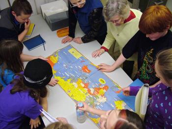 Fourth and fifth grade students from Captain Albert School in Belfast crowd around a geopolitical map of World War II following a presentation at the school by Belfast Museum and Historical Society Director Megan Pinette, top, center. (Photo by Ethan Andrews)