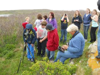 Birder Nancy Bither, seated, plays bird calls on her iPad for students, as Will Perkins sights seabird nests through her telescope. Other students wait their turn while Jack Buthy uses binoculars to observe other birds migrating via Damariscove Island. Courtesy of Rita Arnold